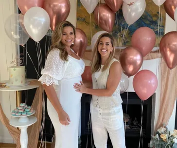 Two women smiling, one pregnant, standing amidst pink and white balloons, with a cake and macarons on a table nearby.