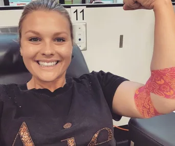 Person smiling and flexing arm with colorful bandage, sitting in a chair, possibly after donating blood.