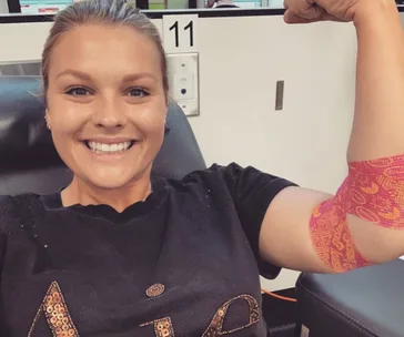 Person smiling and flexing arm with colorful bandage, sitting in a chair, possibly after donating blood.