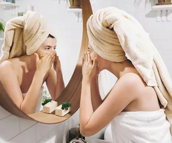 Woman exfoliating face in a bathroom mirror, wearing a towel wrap on her head and body.