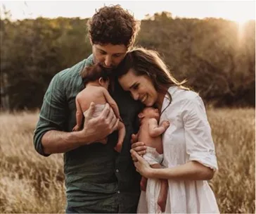 A couple in a field, each holding a newborn, smiling and embracing as the sun sets behind them.