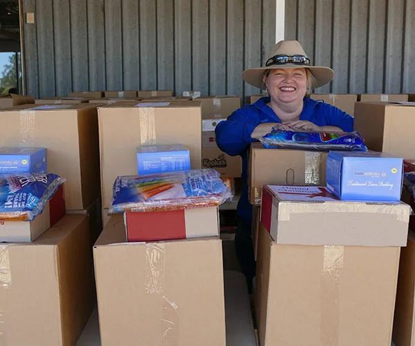 A person in a hat smiling, surrounded by stacks of boxes and supplies, likely part of a relief or support effort.