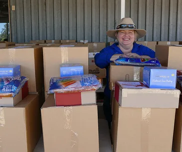 A person in a hat smiling, surrounded by stacks of boxes and supplies, likely part of a relief or support effort.