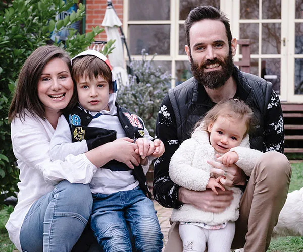 A smiling family of four poses outdoors, with parents holding a young son and daughter in their arms.