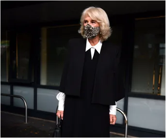 A woman wearing a black outfit with a white-collared shirt and a leopard print mask stands outdoors holding a handbag.