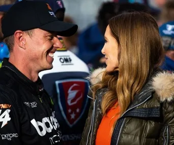 Two people smiling and talking at a motorsport event, one is in racing gear.