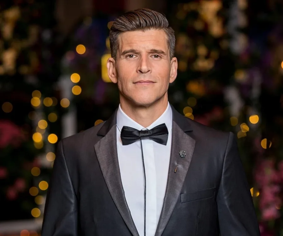 Man in tuxedo with bow tie, standing against a blurred background of colorful lights.