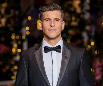 Man in tuxedo with bow tie, standing against a blurred background of colorful lights.