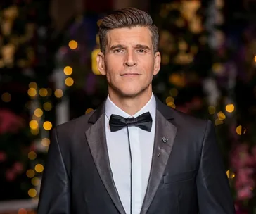 Man in tuxedo with bow tie, standing against a blurred background of colorful lights.
