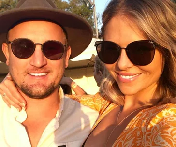 Young couple smiling outdoors; man wearing hat and sunglasses, woman in sunglasses with arm around him.