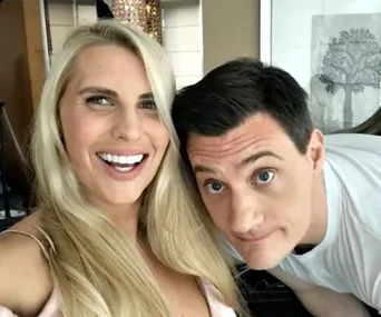 Two people smiling, with long blonde hair on the left and short dark hair on the right, in a casual indoor setting.