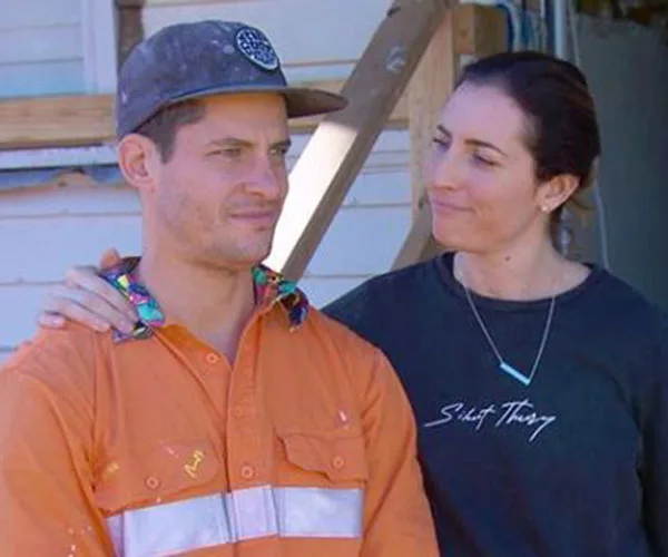 Two people stand together, one wearing an orange shirt and cap, the other in a black sweatshirt, in front of a building site.