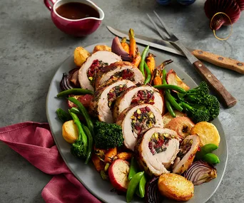 Sliced stuffed turkey roulade with roasted vegetables on a platter, accompanied by gravy.