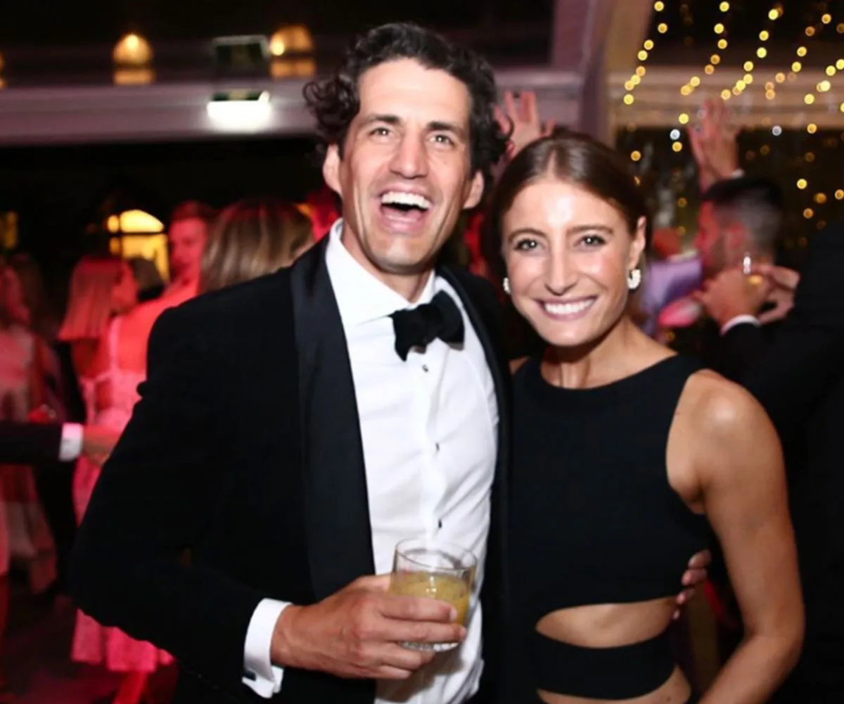 A couple smiling at a formal event, he holds a drink, vibrant lights in the background.