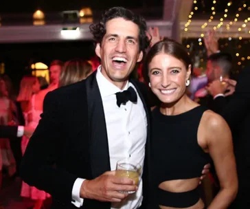 A couple smiling at a formal event, he holds a drink, vibrant lights in the background.