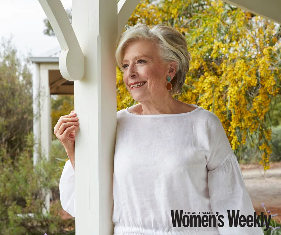 Mature woman in a white dress leans against a porch post, smiling, with yellow flowers in the background.