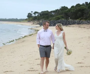 A couple in wedding attire walks hand-in-hand on a sandy beach, with ocean waves and greenery in the background.