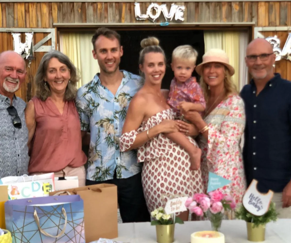 Family smiling at a celebration with gifts on table, child in arms, and "LOVE" sign in background.