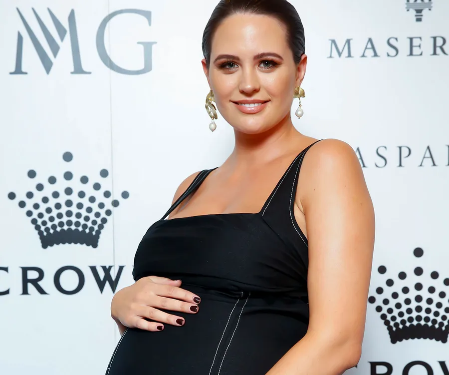 Pregnant woman in black dress, smiling, attending a media event with logos behind her.