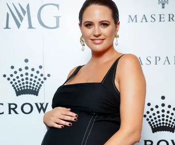 Pregnant woman in black dress, smiling, attending a media event with logos behind her.