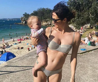 Woman in bikini holding baby at a sunny beach, surrounded by people and vibrant scenery.