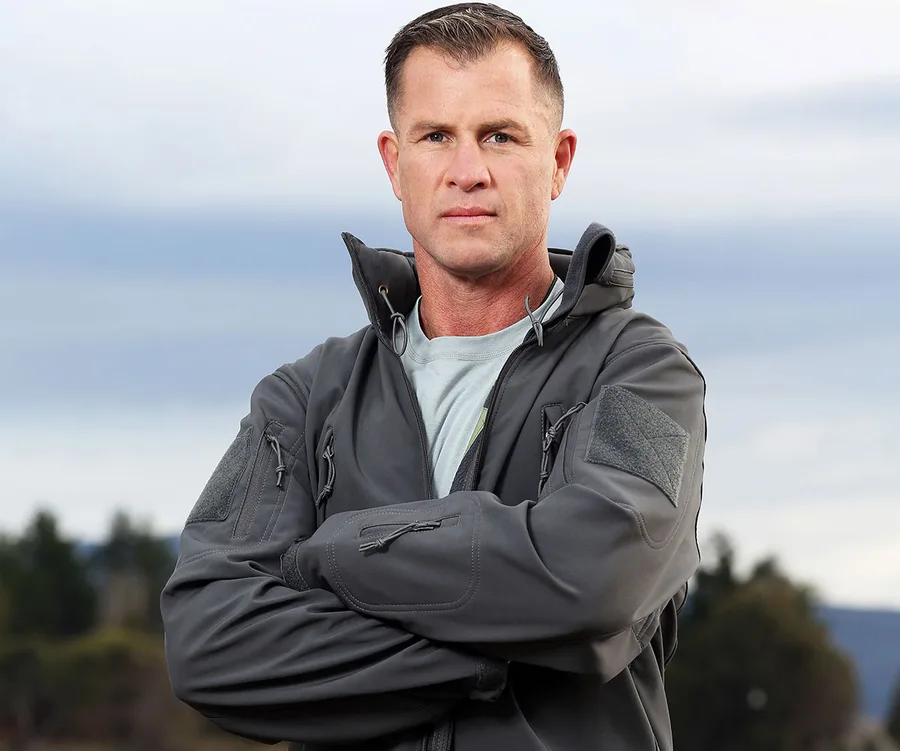 Man in a gray jacket stands outdoors with arms crossed against a cloudy sky background.
