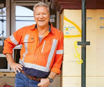 Man in orange high-vis jacket smiling, standing at a construction site beside a marked brick wall, hand on hip.
