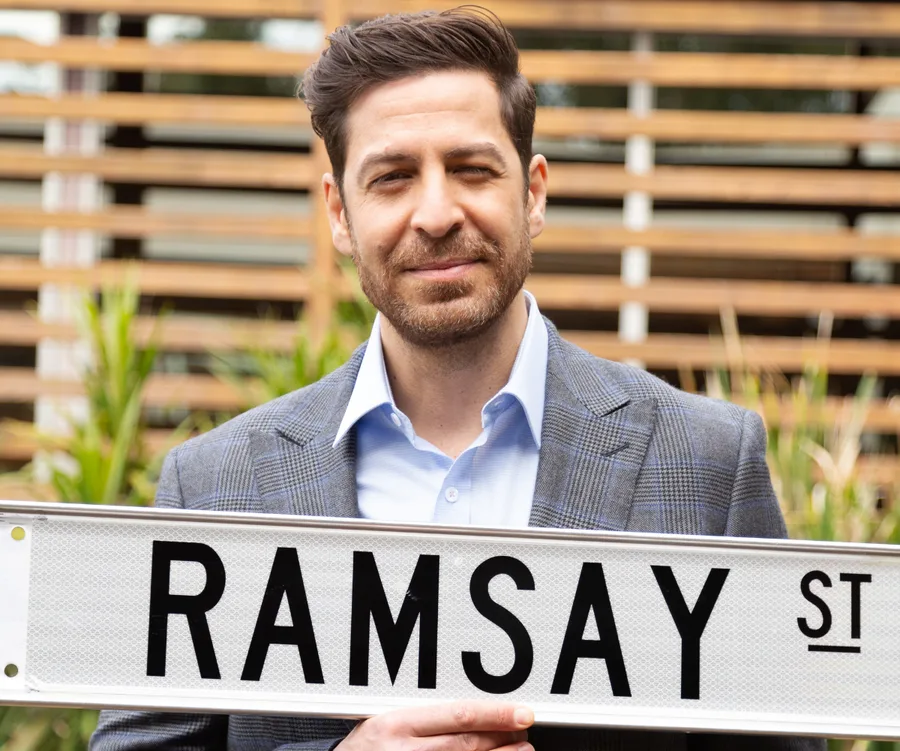 Man in a checkered suit holding a "Ramsay St" sign, standing outdoors with a blurred wooden fence in the background.