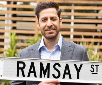 Man in a checkered suit holding a "Ramsay St" sign, standing outdoors with a blurred wooden fence in the background.