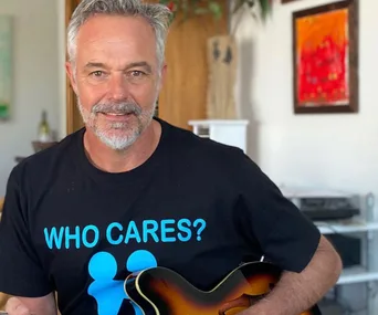 Man with gray beard, wearing black "WHO CARES?" shirt, holds a guitar indoors with colorful art in the background.