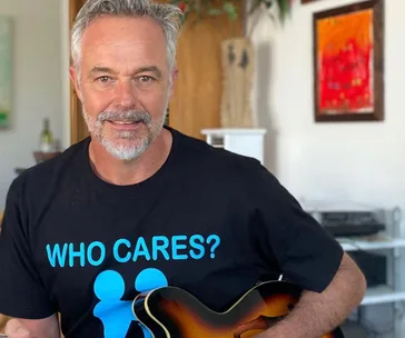 Man with gray beard, wearing black "WHO CARES?" shirt, holds a guitar indoors with colorful art in the background.