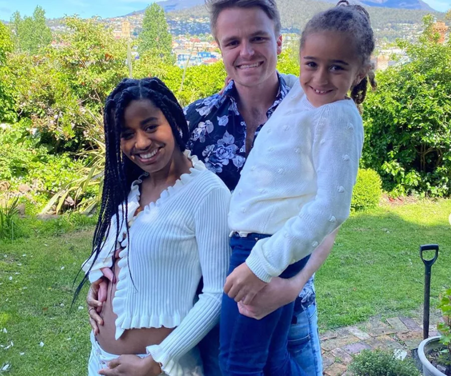 Family smiling together outdoors, a woman cradles her belly, a man holds a child in a garden setting.