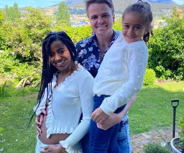 Family smiling together outdoors, a woman cradles her belly, a man holds a child in a garden setting.