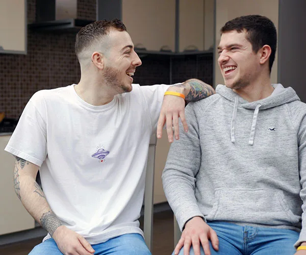 Two smiling young men sitting, one in a white T-shirt, the other in a gray hoodie, with an arm around each other.