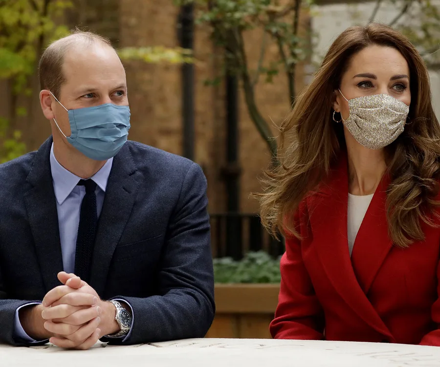 Two people sitting at a table, wearing masks, one in a suit with a blue mask and the other in a red coat with a floral mask.