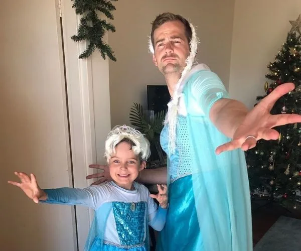 Father and son dressed as Elsa from Frozen, posing dramatically in a living room with a Christmas tree.