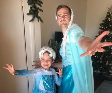 Father and son dressed as Elsa from Frozen, posing dramatically in a living room with a Christmas tree.