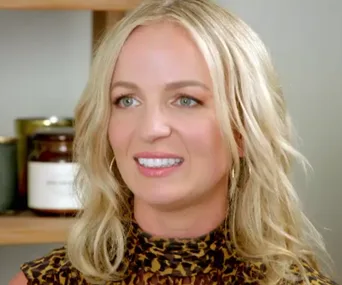 Blonde woman with long hair smiles, wearing a leopard print top, with jars in the background.