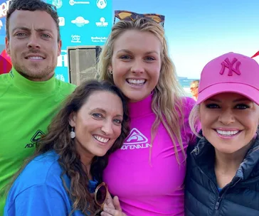 Four people smiling at a beach event, wearing colorful shirts and a pink cap, with a blue sky backdrop.
