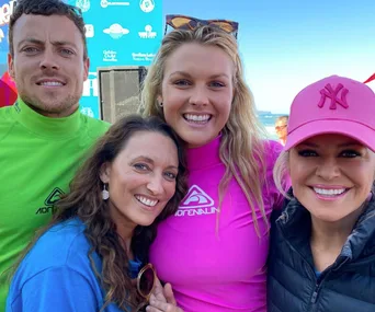 Four people smiling at a beach event, wearing colorful shirts and a pink cap, with a blue sky backdrop.