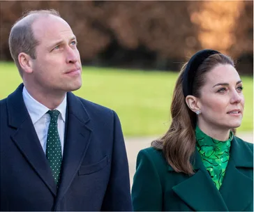Two individuals, one in a dark coat and polka-dot tie, the other in a green outfit, looking upwards outside.