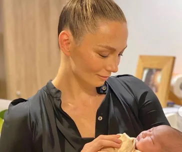 Woman with hair in a bun gently holding and looking at an infant wrapped in a blanket indoors.