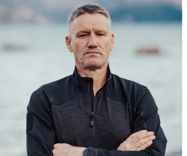 Man in a black jacket standing with arms crossed outdoors by a lake.