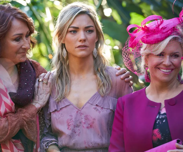 Three women at an outdoor event; one in pink lace dress with pink fascinator, another in blush dress with supportive gesture.