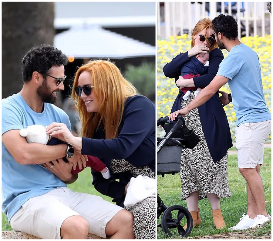 Couple smiling and holding a baby outdoors, near a stroller in a park setting.