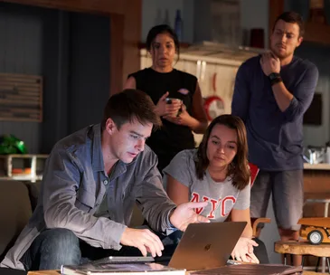 Group of four people focused on a laptop screen in a dimly lit room, suggesting a serious discussion or investigation.