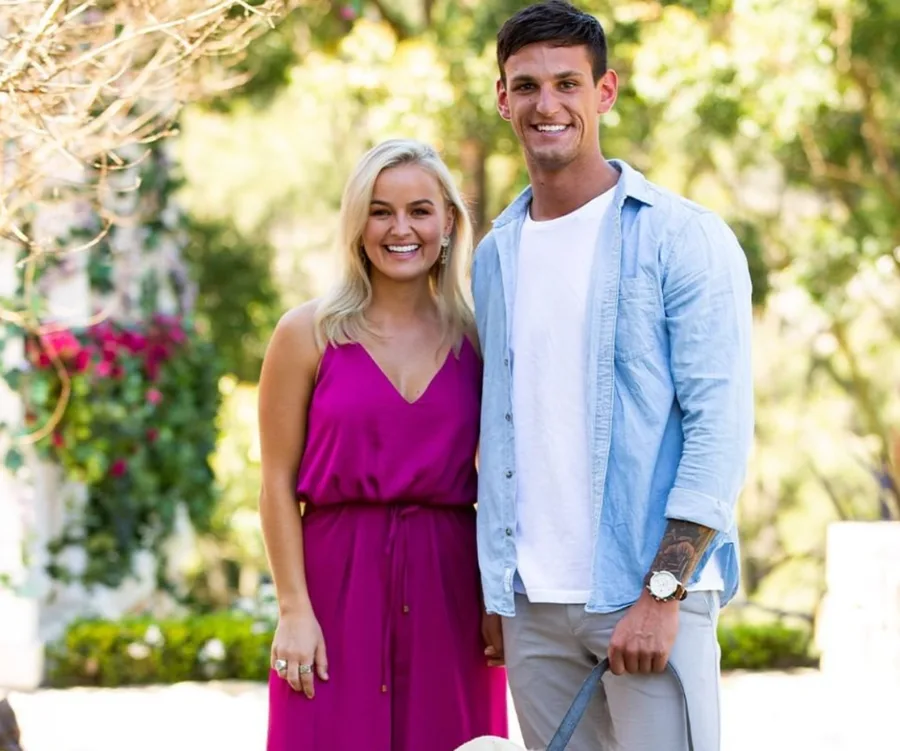 A couple standing outdoors, smiling at the camera, with greenery in the background.