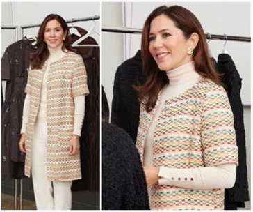 A woman in a colorful patterned coat over a white turtleneck, standing near clothing racks, smiling.