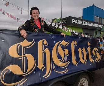 Shelvis impersonator in black outfit leaning on sign at outdoor event, with ornate "Shelvis" text and decorations.
