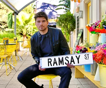 Man in black jacket holds "Ramsay St" sign, sitting at an outdoor cafe with yellow chairs and colorful flowers.